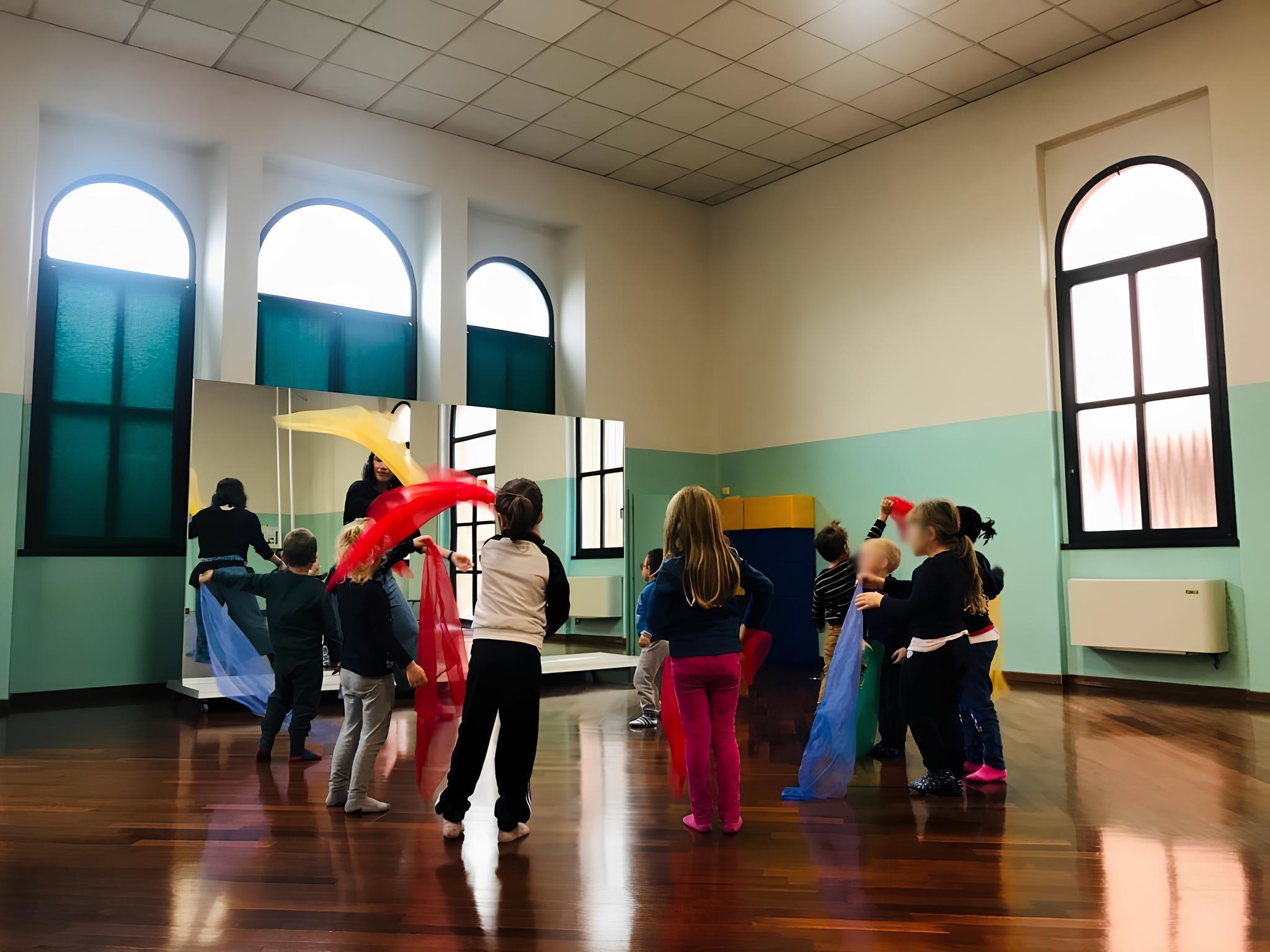 A group of preschool children playing with colorful scarves in a classroom with a wooden floor and large windows