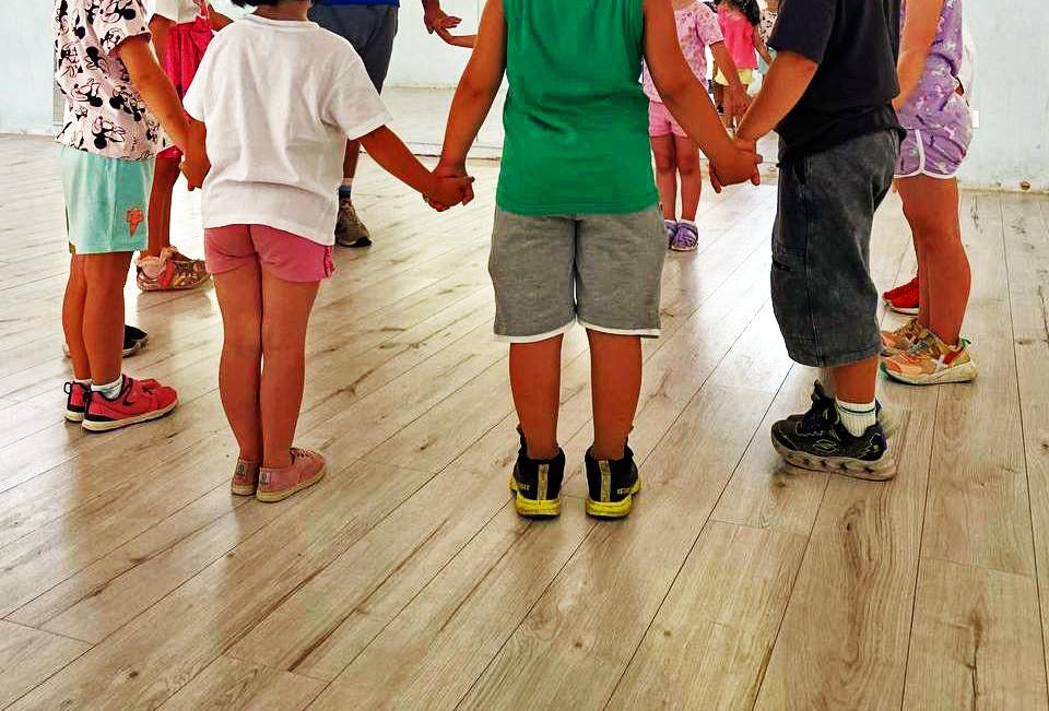 a group of young children standing in a circle, holding hands. They are indoors on a wooden floor, dressed in casual summer clothing, with sneakers of various colors. The children are standing close together, suggesting a sense of unity and friendship. The photo captures the scene from waist-down, focusing on the children's legs and feet, and the different shades and textures of their clothing and shoes. 