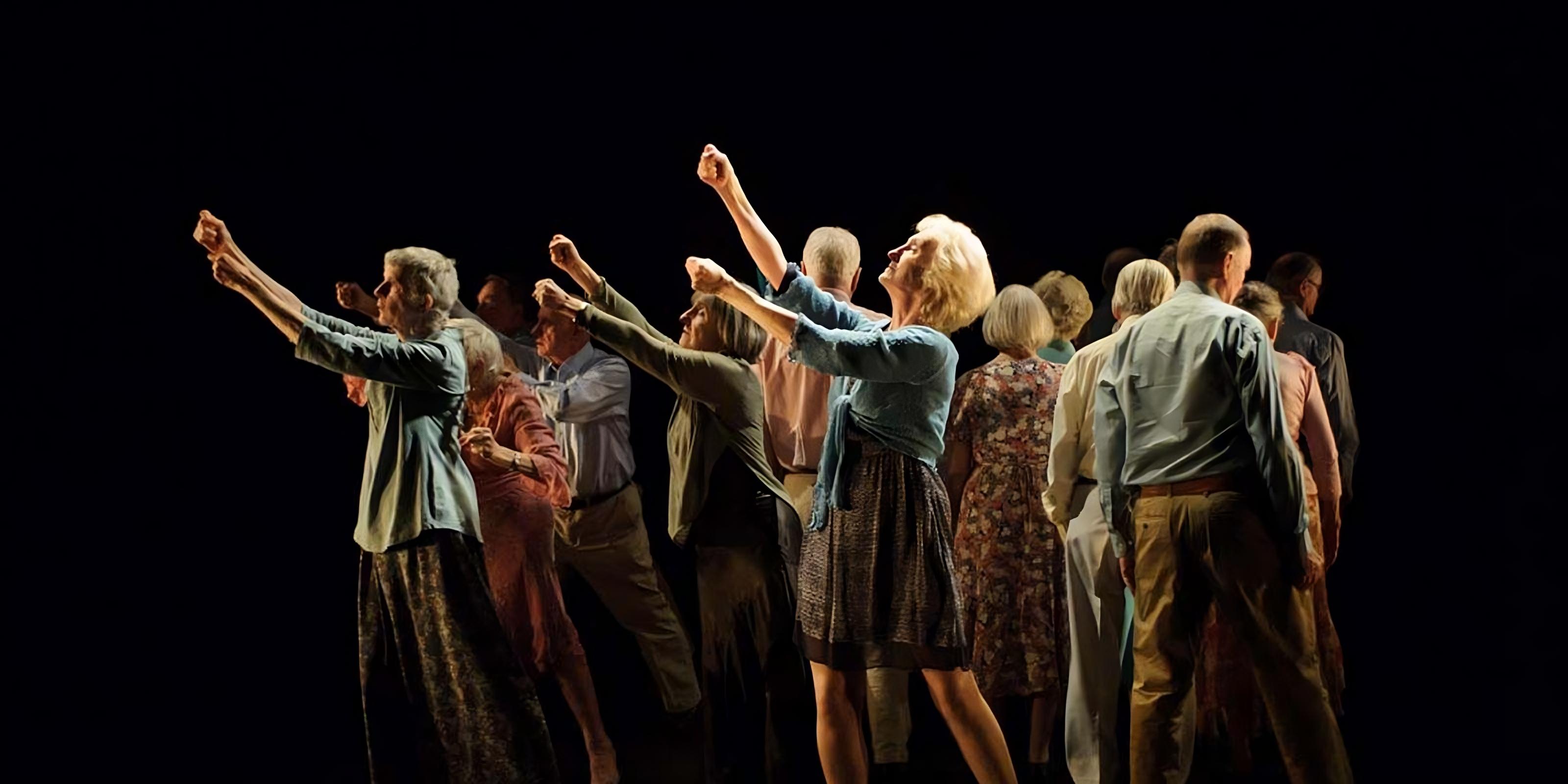 A group of older adults engaged in a dance or performance. They are positioned closely together, facing different directions, with some reaching their arms upward in expressive gestures. The group is dressed in comfortable, casual attire, and the lighting is focused on them, highlighting their movements against a dark background, creating a dramatic and intimate atmosphere.